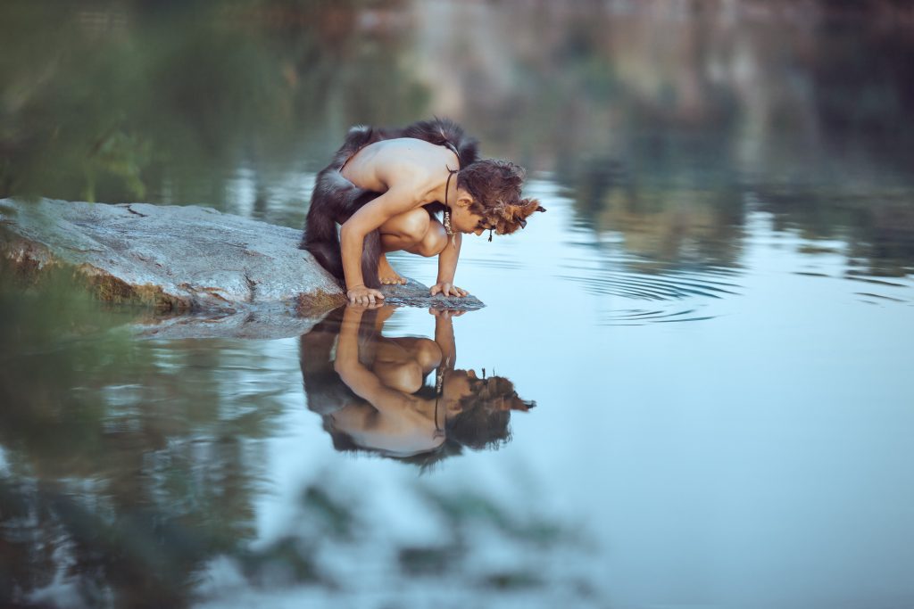 Caveman boy sitting on the rock and looking at him self in the water reflection in lake.